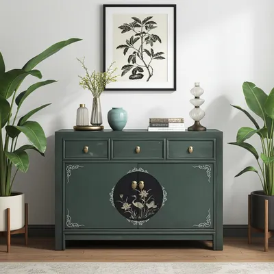 A green sideboard with decorative elements, flanked by potted plants, with artwork and a lamp on top.
