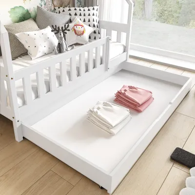 A white bed with a pull-out trundle bed containing folded clothes, set against a window with a view of trees.