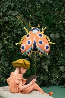 A child is sitting on a cushion, reading a book, with a large ladybug balloon floating behind them against a backdrop of green foliage and fairy lights.
