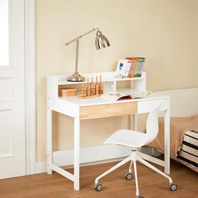 A white desk with a wooden drawer front, a silver desk lamp, and some books on a shelf.