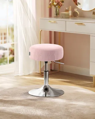 A pink velvet stool with a silver adjustable base is placed on a light beige rug in front of a white vanity desk with gold accents.