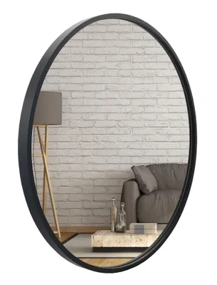 A round mirror with a black frame is shown against a white brick wall background. In the reflection, a floor lamp, a sofa, and a coffee table with decorative bottles are visible.
