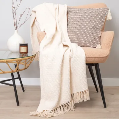 A cream-colored throw blanket is draped over a chair next to a small table with a vase and a candle.