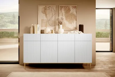 A white sideboard with gold legs is displayed in a modern living room setting, with artwork on the wall and large windows overlooking a landscape.