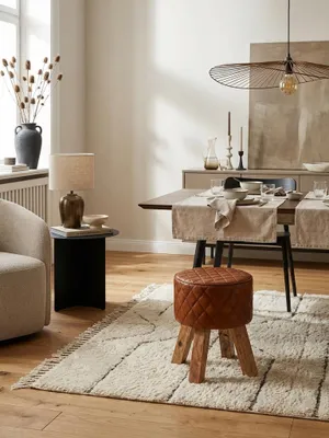 Eleganter Sitzhocker aus braunem Echtleder und Mango-Holz in einem hellen Wohnzimmer, ideal als bequeme Sitzgelegenheit neben einem Sofa auf einem weichen Teppich._#sku_SAFWL6.090#