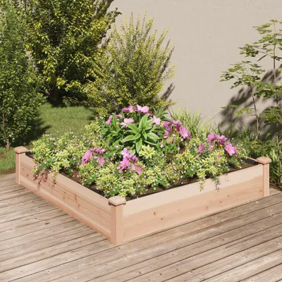 A raised garden bed filled with plants and flowers sits on a wooden deck, with trees and a wall in the background.