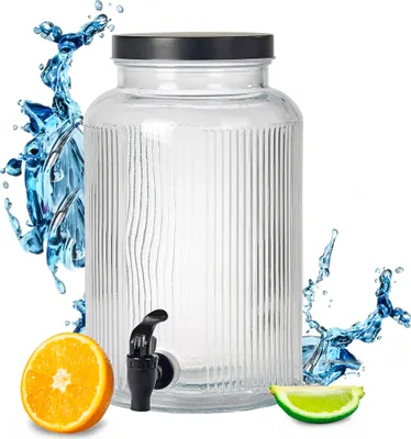 A glass beverage dispenser with a black lid and a black spigot, surrounded by splashes of water and slices of orange and lime.