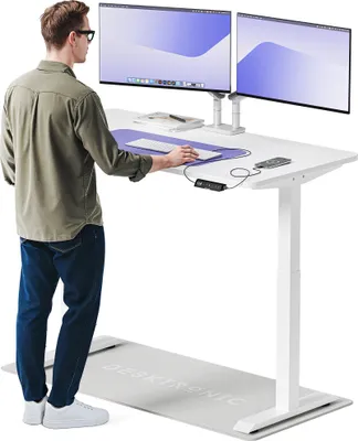 A man is standing at a white standing desk with two monitors, using a laptop. There is an anti-fatigue mat on the floor in front of the desk.