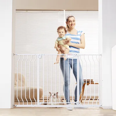 A woman holds a baby while standing behind a white baby gate. A cat is visible on the floor behind the gate.