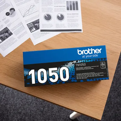A Brother TN-1050 toner cartridge box is placed on a wooden desk next to some documents.