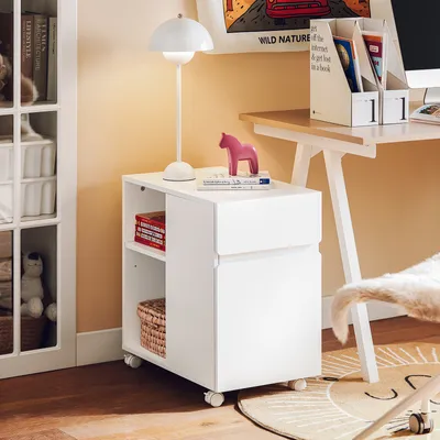 A white rolling cabinet with shelves and drawers, next to a desk with a lamp and books.