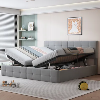 A gray upholstered bed frame with the mattress lifted, revealing storage space underneath. The bed has a tufted headboard and is positioned in a room with blinds, shelves, and artwork.
