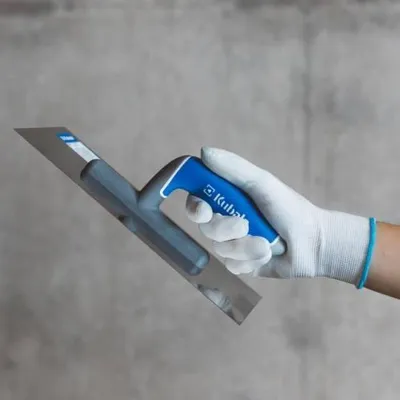 A hand wearing a white glove holds a trowel against a gray concrete wall.