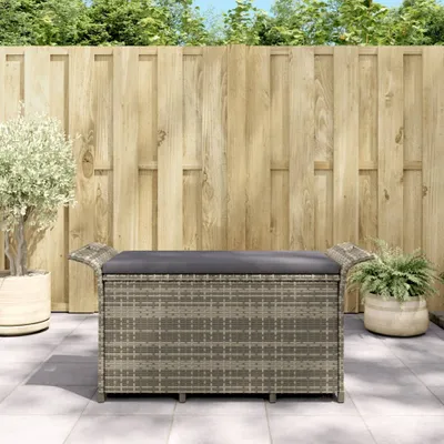 A rattan storage bench with a grey cushion sits on a patio in front of a wooden fence.