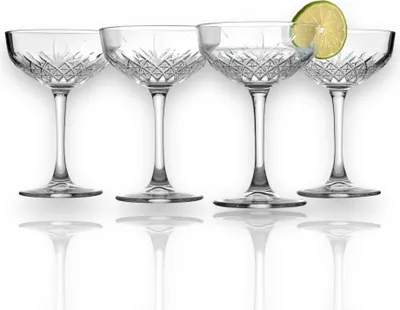 Four crystal champagne coupes are arranged in a row on a white background, with reflections visible below them. One of the coupes contains a clear liquid and is garnished with a slice of lime.