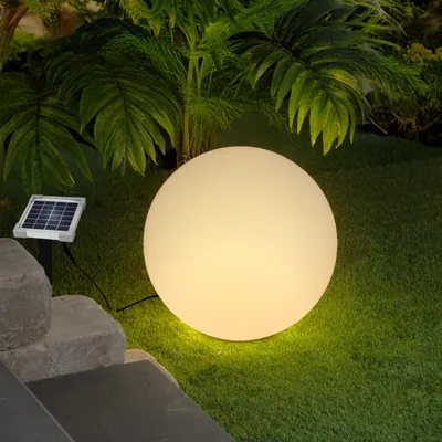 A solar-powered decorative sphere light sits on a lawn next to some stone steps, with lush green foliage in the background.