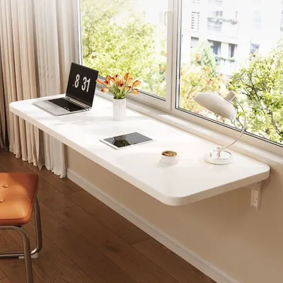 A white folding desk mounted on a wall next to a window, with a laptop, tablet, lamp, and coffee cup on it.