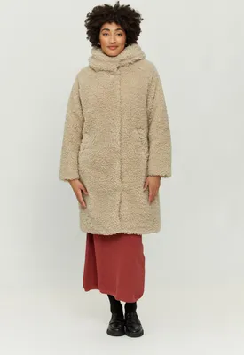 A woman is standing and posing in a studio setting, wearing a beige teddy coat over a red dress.