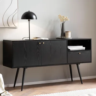 A black wooden sideboard with golden handles on legs, decorated with a lamp, books, and a vase with dried flowers.