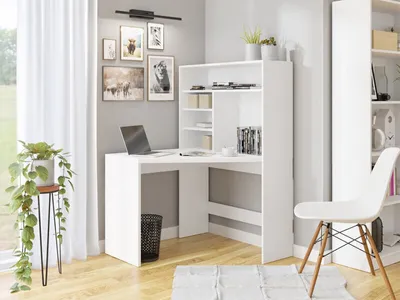 A white corner desk with shelves is set up in a room with a window and curtains. There are framed pictures on the wall and a chair next to the desk.