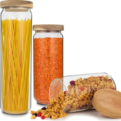 Three glass jars with wooden lids filled with spaghetti, lentils, and granola, with a wooden spoon and spilled granola.
