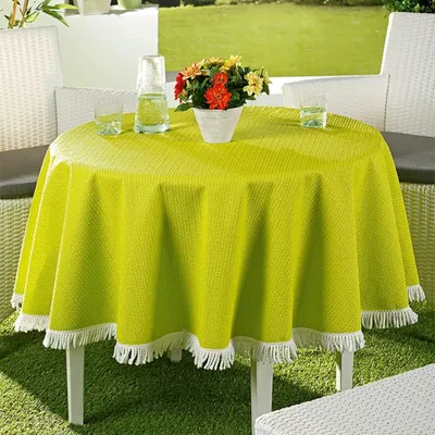 A round green tablecloth with white fringe is set on an outdoor table on a patio with chairs and greenery in the background.