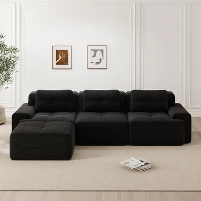 A black modular L-shaped sofa with an ottoman is shown in a well-lit room with white walls and decorative molding. Two framed abstract art pieces hang on the wall above the sofa. A plant is partially visible on the left, and a book lies open on the rug in 