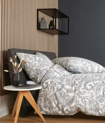 A bed with patterned bedding and pillows, next to a small wooden side table with a vase of dried flowers and a laptop.