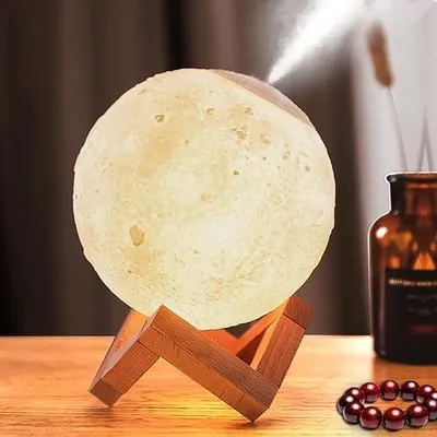 A moon-shaped humidifier and night light sits on a wooden table, emitting a mist. A brown glass bottle and a string of red beads are also visible.