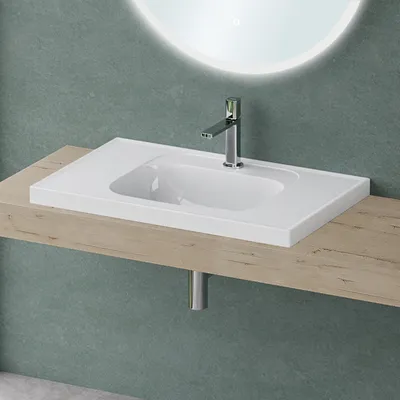 A white rectangular sink with a chrome faucet is mounted on a wooden shelf against a textured green wall. A circular mirror with a light rim is visible in the background.