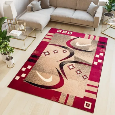 A modern rug with a geometric and abstract design is placed on a light wooden floor in a living room setting. A sectional sofa and a glass coffee table are visible in the background.