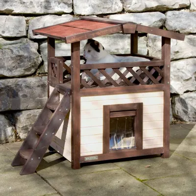 A cat is sitting on a multi-level wooden cat house with a red roof, placed outdoors on a stone patio with a stone wall in the background.