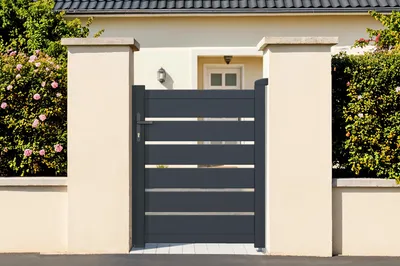 A modern dark gray aluminum garden gate is shown in front of a light-colored house with green bushes on either side.