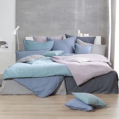 A bed with stone-washed bedding in various shades of pink, blue, and gray, set against a textured gray wall.