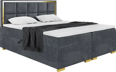 A dark gray upholstered boxspring bed with a gold frame around the headboard, two white pillows, and a dark gray blanket draped over the mattress.
