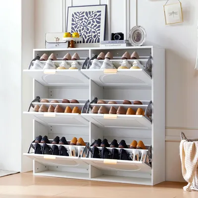 A white shoe cabinet with multiple shelves and flip-down doors, filled with shoes, placed in a room with a framed picture and a camera on top.