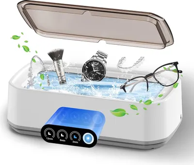 A white ultrasonic cleaner with a transparent lid, filled with water and cleaning various items like glasses, a watch, jewelry, and a makeup brush. There are green leaf graphics around the cleaner.