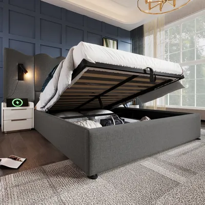 A bedroom scene with a bed that has its storage compartment open, revealing stored items. A bedside table with a lamp is also visible.