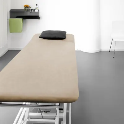 A treatment table with a taupe cover and a black pillow, in a clean, bright room with a shelf holding a green towel and some bottles.