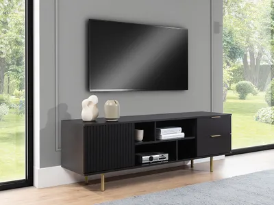 A modern living room with a TV mounted on the wall above a black TV stand with gold legs. The TV stand has a fluted door, drawers, and open shelves. A large window with a view of a garden is on the right.