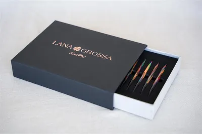 A dark gray box with the Lana Grossa logo and text is partially open, revealing a set of colorful knitting needles inside.