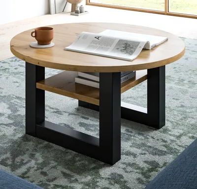A round coffee table with a wooden top and black metal legs, placed on a patterned rug in a living room setting. A magazine and a cup of coffee are on the table.