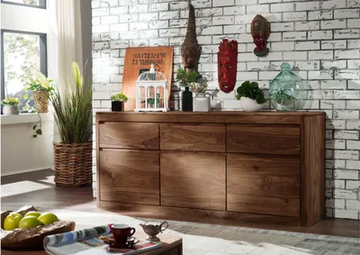 A wooden sideboard is displayed in a living room setting, decorated with various items on top and a brick wall in the background.