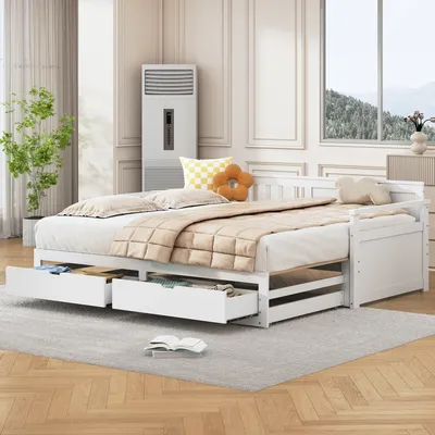 A white wooden daybed with two drawers pulled out, set up in a bedroom with a plant and a window in the background.