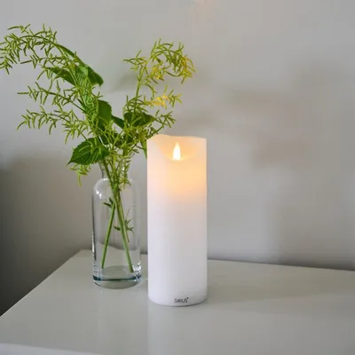 A white LED candle and a vase with green foliage sit on a white surface against a light gray wall.