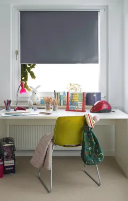 A desk with a roller blind, a chair with bags and a blanket, and various school supplies.