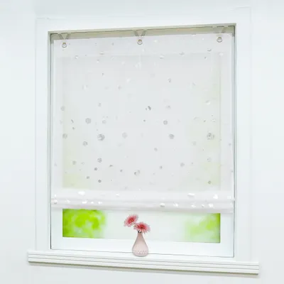 A white roller blind with a circular pattern is shown hanging in a window. A small pink vase with two pink flowers sits on the windowsill below.