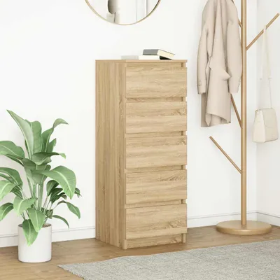 A tall, narrow chest of drawers with five drawers, made of wood-effect material, stands against a white wall. A large green plant in a white pot is to the left, and a coat rack with a beige coat and a white handbag is to the right. A round mirror is visibl