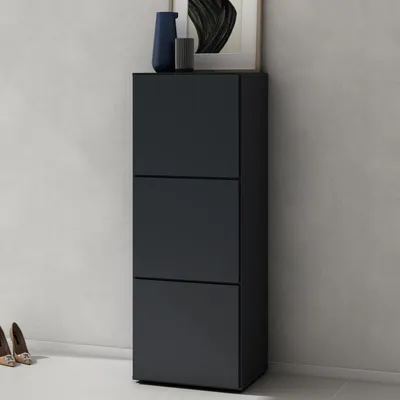 A dark gray shoe cabinet with three drawers stands against a light gray wall. Two pairs of women's shoes are visible on the floor to the left of the cabinet. A framed picture and two decorative vases are on top of the cabinet.