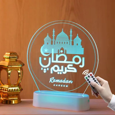 A Ramadan-themed LED lamp with Arabic calligraphy and a mosque silhouette is displayed on a table next to a golden lantern. A hand is holding a remote control, suggesting the lamp is adjustable.